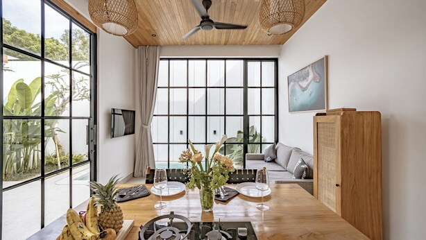 Een moderne eethoek met een houten tafel gedekt voor twee personen, een plafondventilator, grote ramen en uitzicht op groen buiten.
