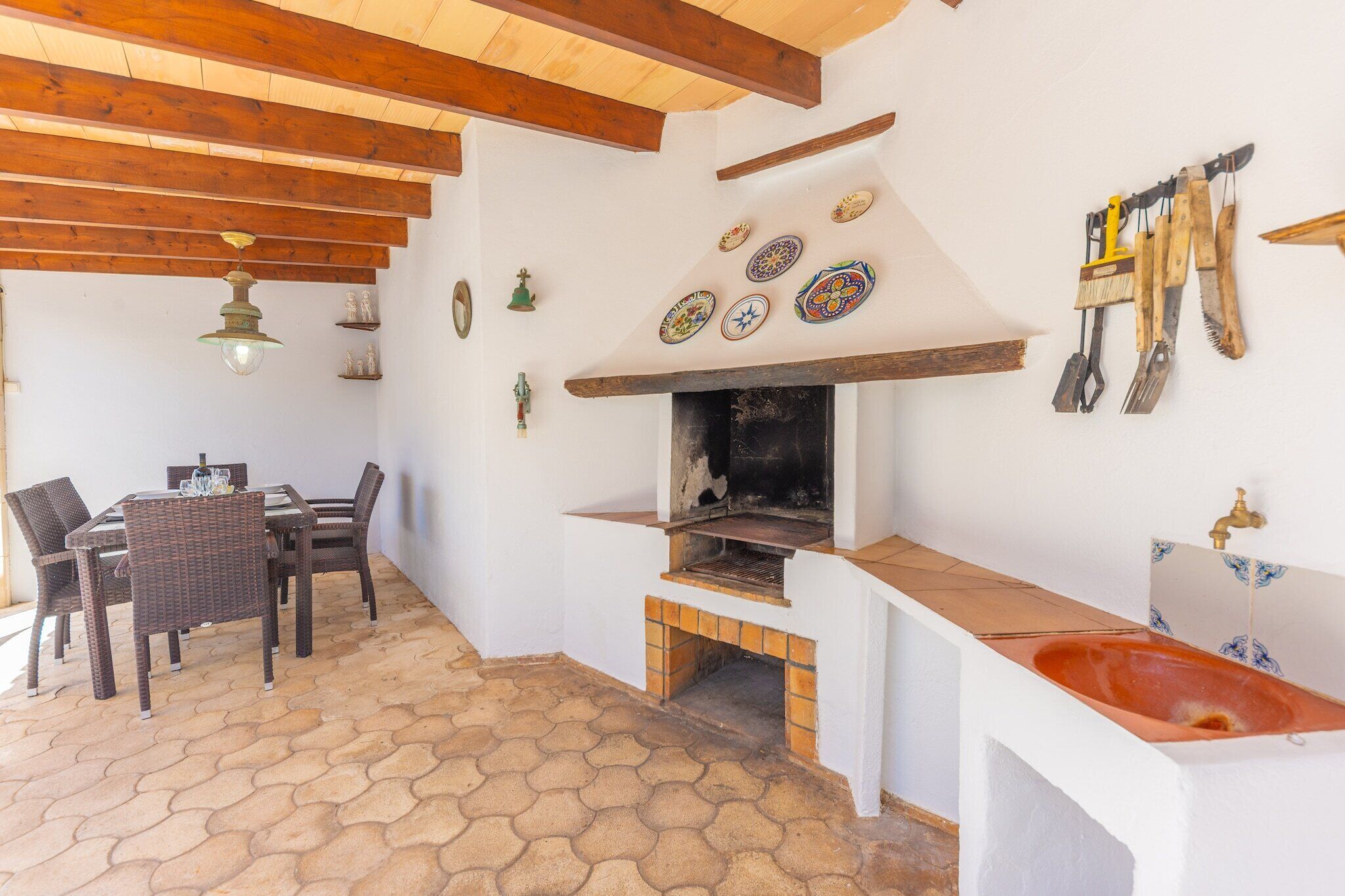A rustic outdoor kitchen with a brick oven, a dining table with chairs, and a hanging set of kitchen utensils.