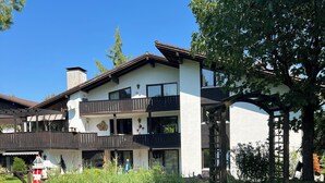 Un edificio a due piani con balconi, un'area giardino con una panchina di legno e un cielo azzurro intenso.