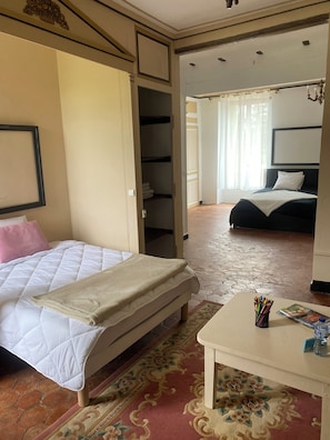 Ein Schlafzimmer mit einem Bett, einem Nachttisch mit Zeitschrift und Wasserflasche, einem Kleiderschrank und Blick auf einen weiteren Raum mit einem Bett.
