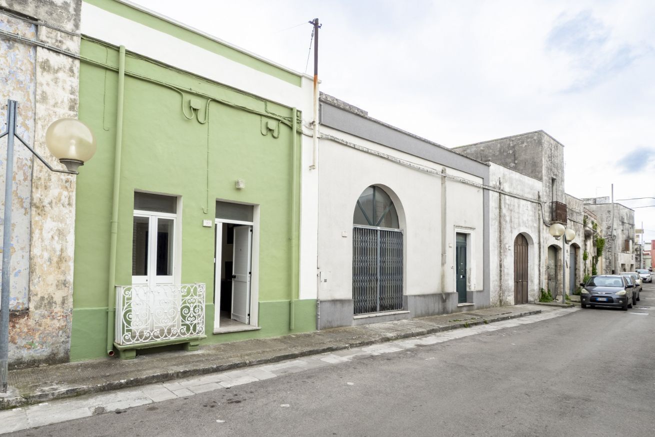 A street with buildings, one with a green facade and a white door, another with a large arched window, and a parked car.