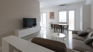 Ein modernes Wohnzimmer mit Flachbildfernseher, einem Essbereich mit Tisch und Stühlen sowie einer Couch mit Kissen.