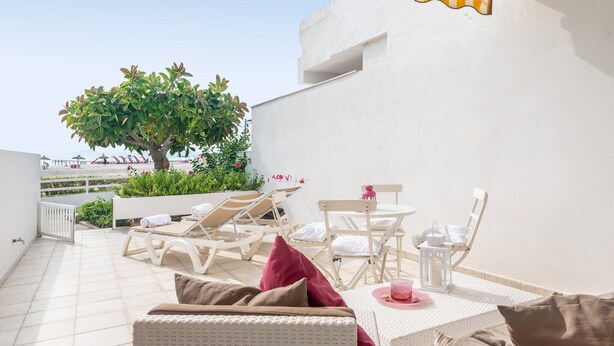 A rooftop terrace with white wicker furniture, a dining table with a pink candle, and a view of a tree and beach in the background. - Apartamento 23 Edificio Canopus - Apartment (Port D'alcúdia, Illes Balears)