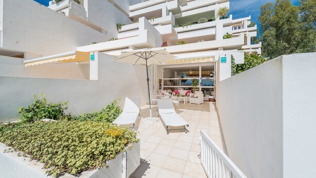 A modern building with white exterior walls, a covered outdoor seating area with lounge chairs, and a view of the sea.