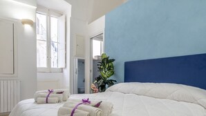 Camera d'albergo con un letto grande, due asciugamani arrotolati con nastri viola, una pianta in vaso e vista su un edificio attraverso una finestra.