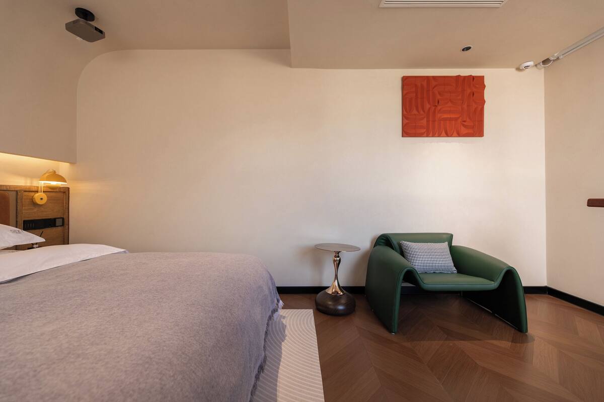 Quarto de hotel moderno com cama bem arrumada, uma poltrona verde, uma mesa pequena e uma obra de arte pendurada na parede.