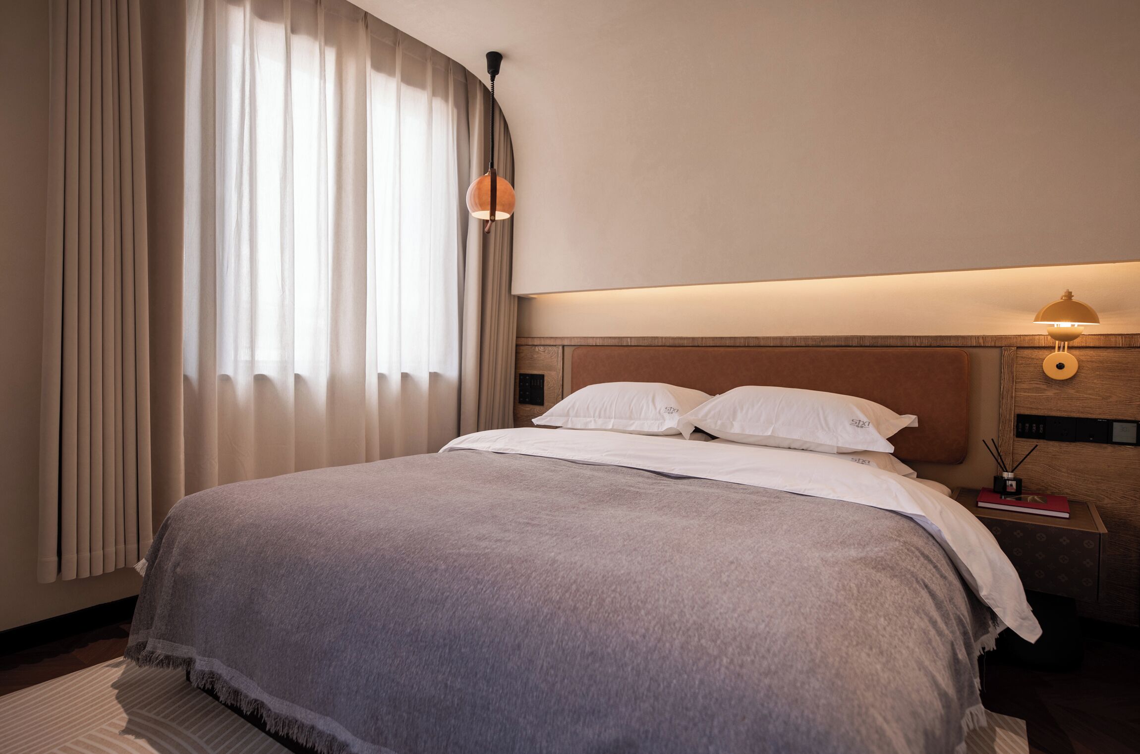 Una cama bien hecha con una manta gris, una mesita de noche con una lámpara y una ventana con cortinas traslúcidas.