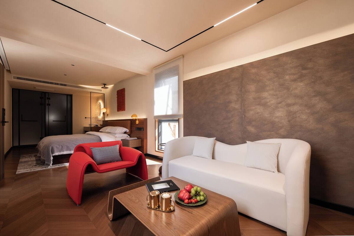 Quarto de hotel moderno com cama, uma poltrona vermelha, sofá branco, mesa de centro de madeira com uma fruteira e uma janela grande.
