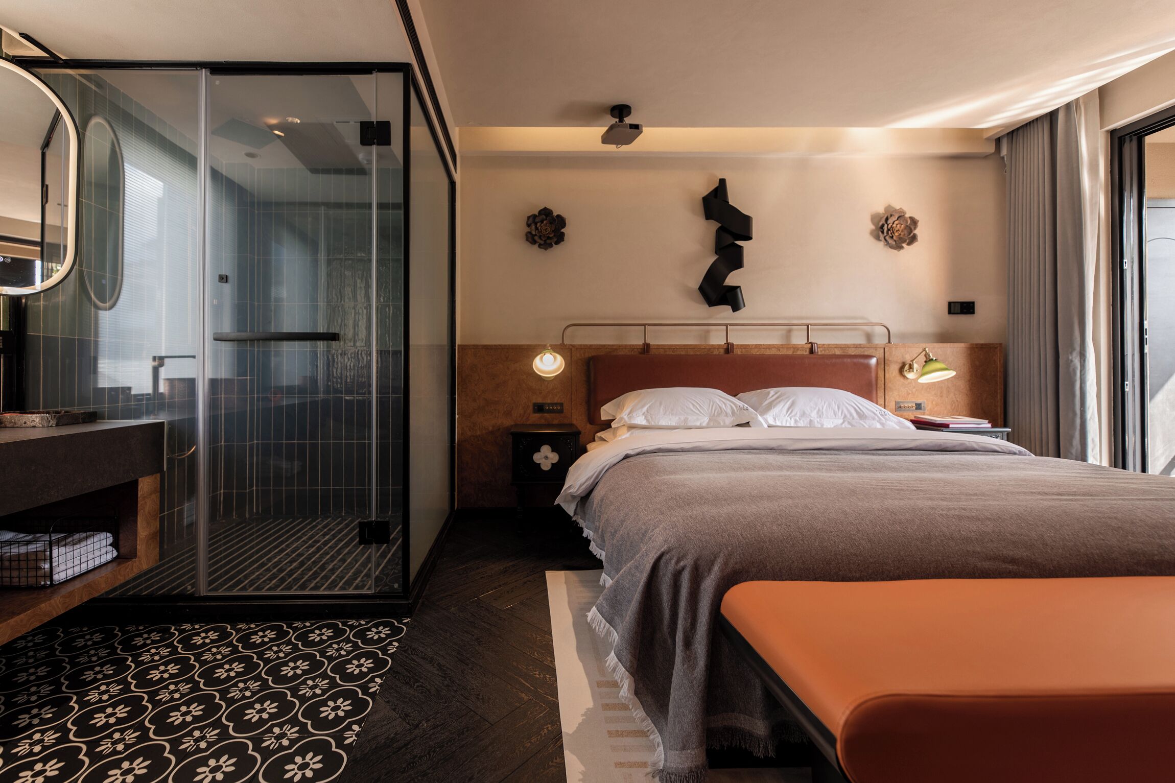 Une chambre d’hôtel moderne dotée d’une douche en verre, d’un banc en bois, d’un lit avec une tête de lit et d’une table de chevet avec une lampe.