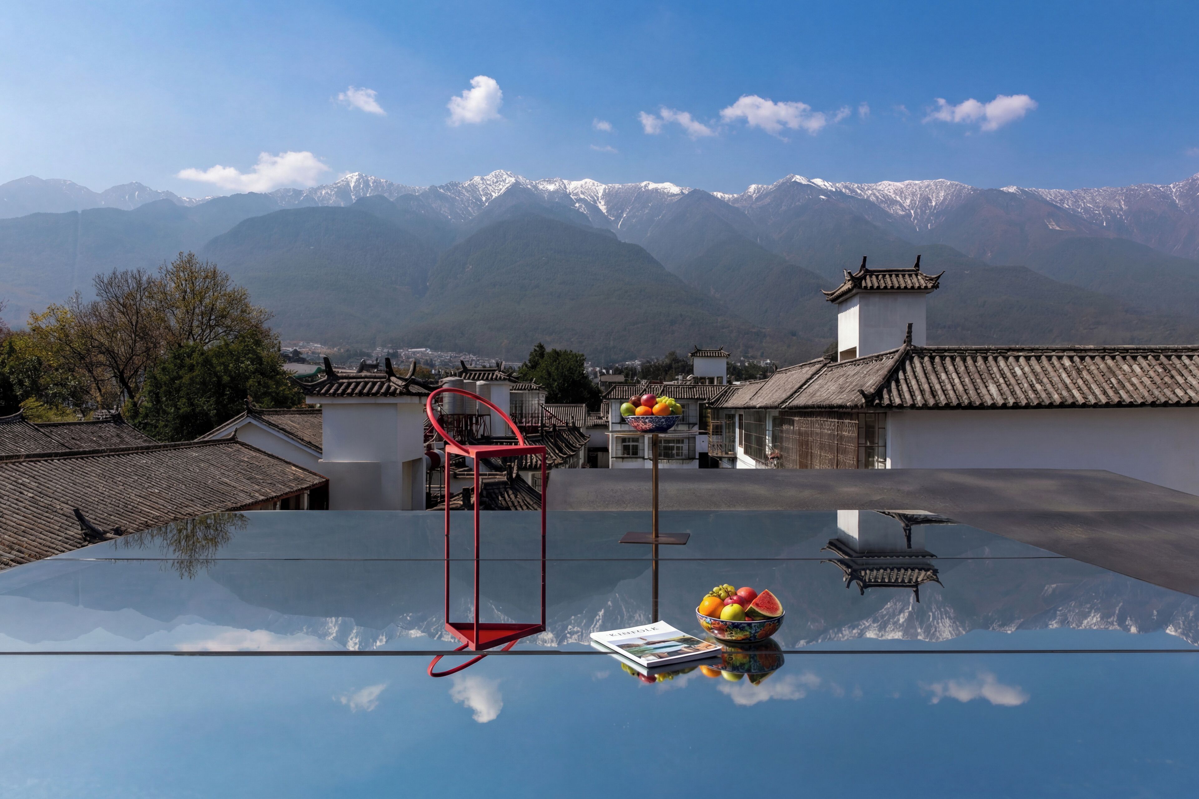 Une chambre d’hôtel moderne avec une table en verre, une chaise rouge et un saladier rempli de fruits.