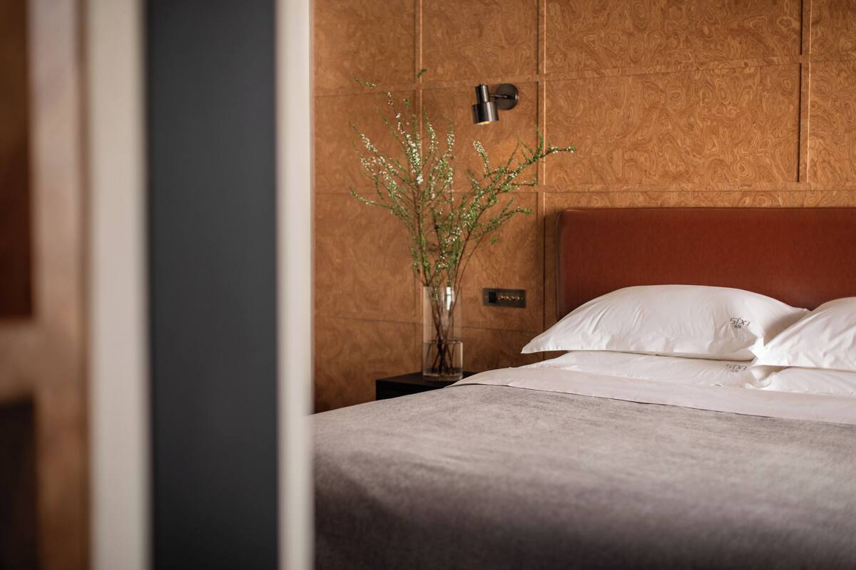 Cama bem arrumada com lençóis brancos, um vaso com folhagem e um luminário fixado na parede.