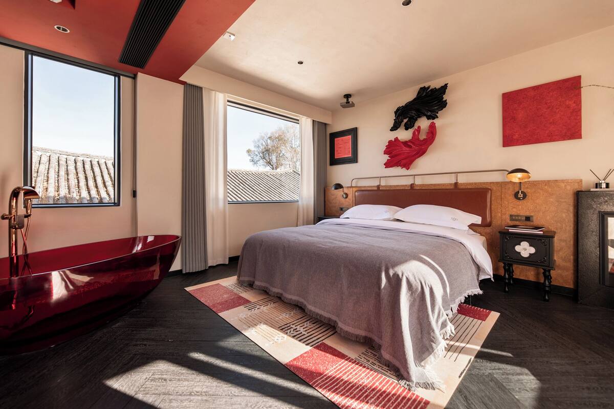 Quarto moderno com uma cama grande, uma banheira vermelha e vista para a paisagem urbana.