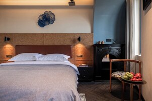 Una cama bien hecha con cabecera, dos lámparas de noche, un adorno en la pared, una mesita de noche de madera oscura con un libro y un pequeño recipiente, y una silla con un frutero.