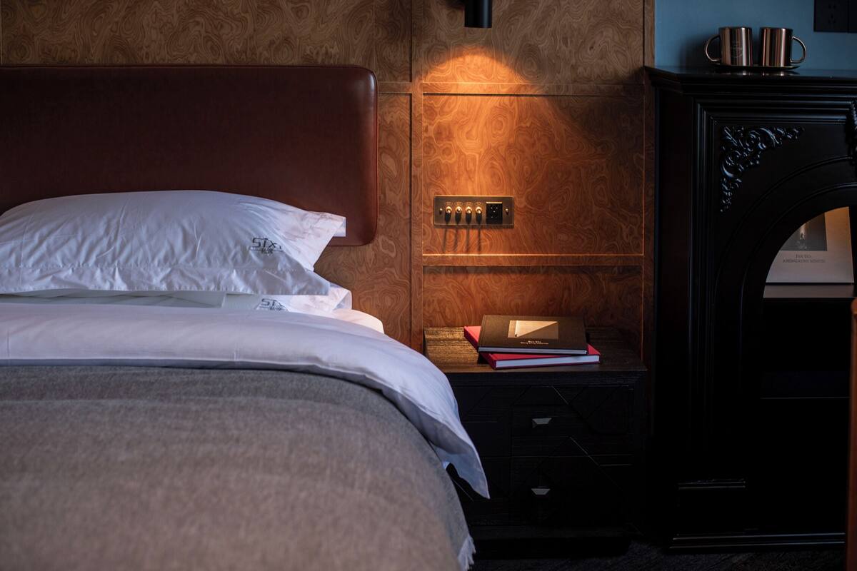 Uma cama bem arrumada com lençóis brancos, cabeceira de madeira, mesa de cabeceira com um livro e uma luminária, e um acessório de luz fixado na parede.