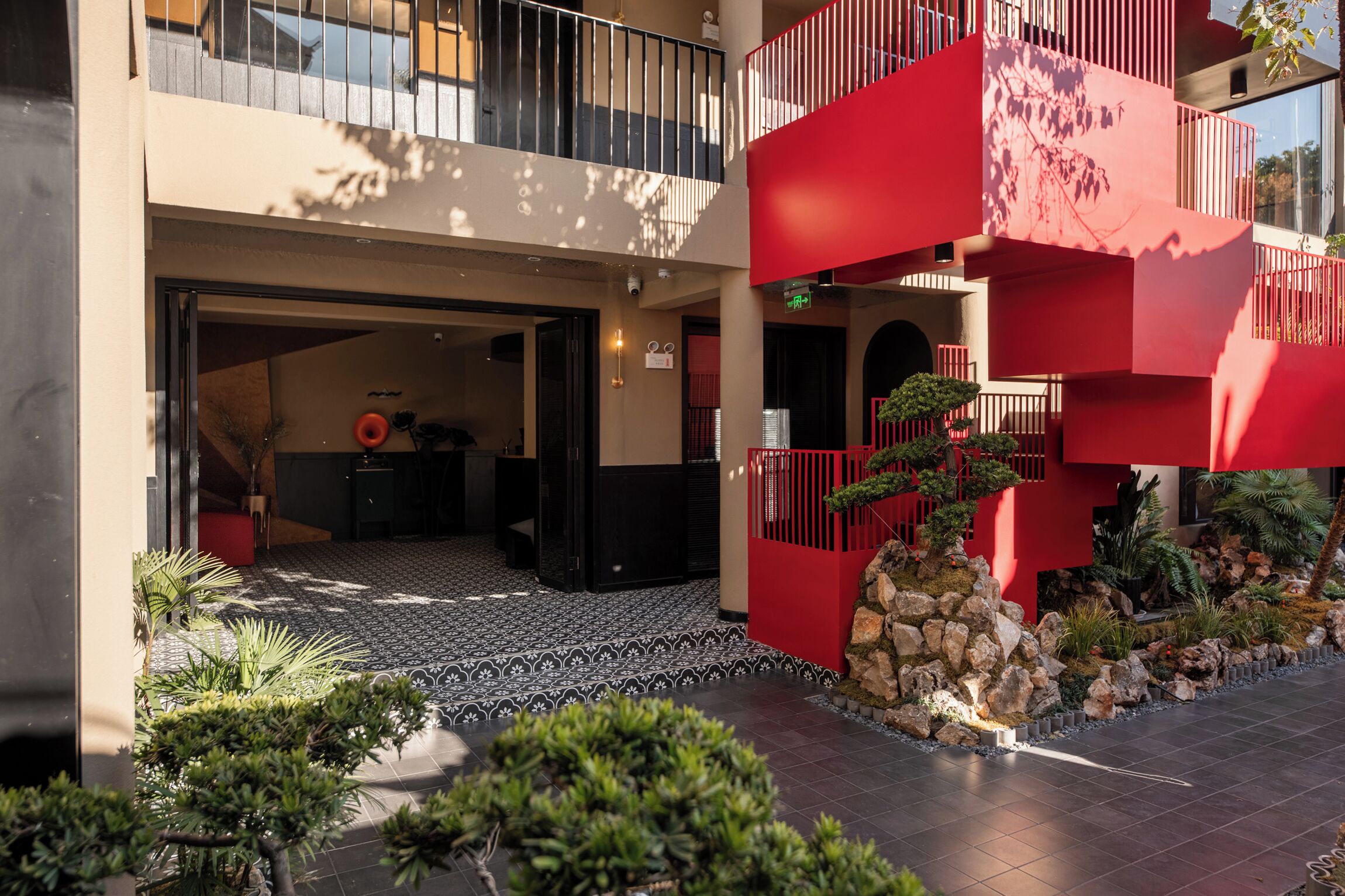 Entrada de hotel con fachada roja y beige, zona de acceso empedrada y un pequeño jardín con rocas y plantas.