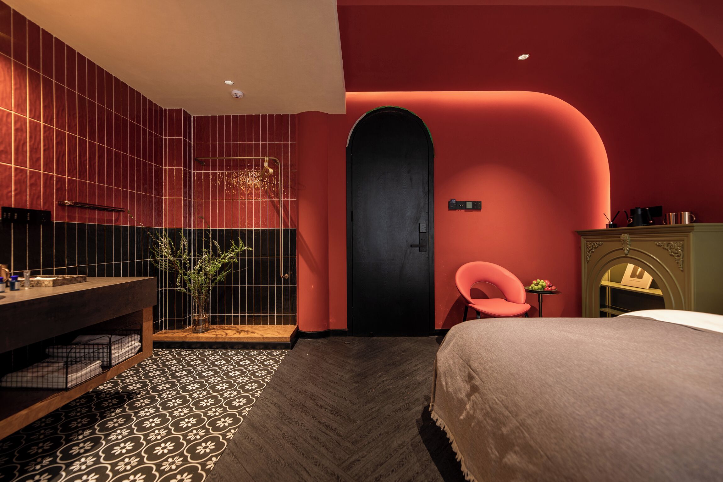 Ein modernes Hotelzimmer mit einer Wand aus roten und schwarzen Fliesen, einem hölzernen Waschtisch, einem Bett mit weißer Daunendecke und einem kleinen Tisch mit einer Pflanze.