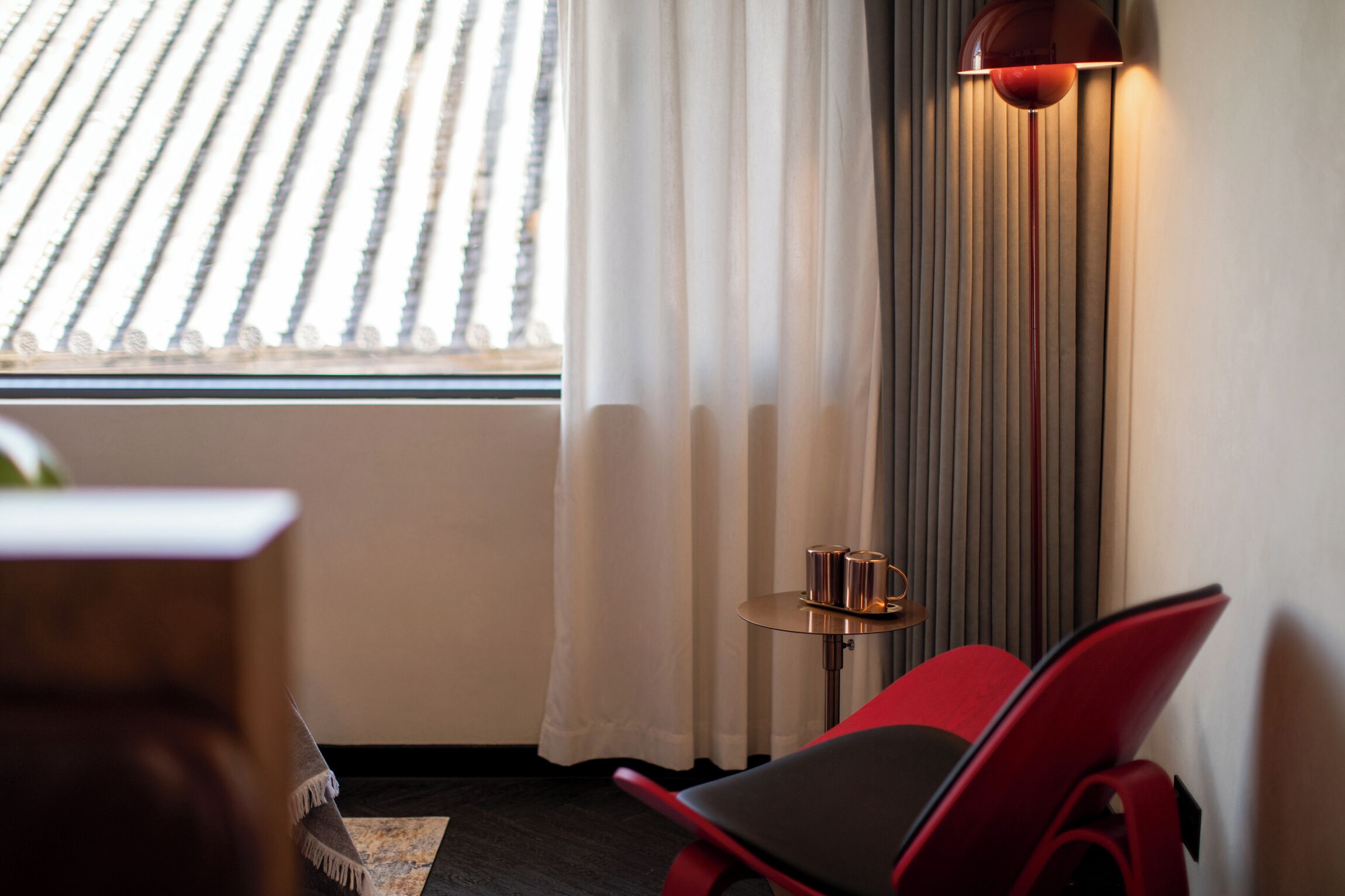 A hotel room with a red chair, a small table with a lamp, and a window with blinds.