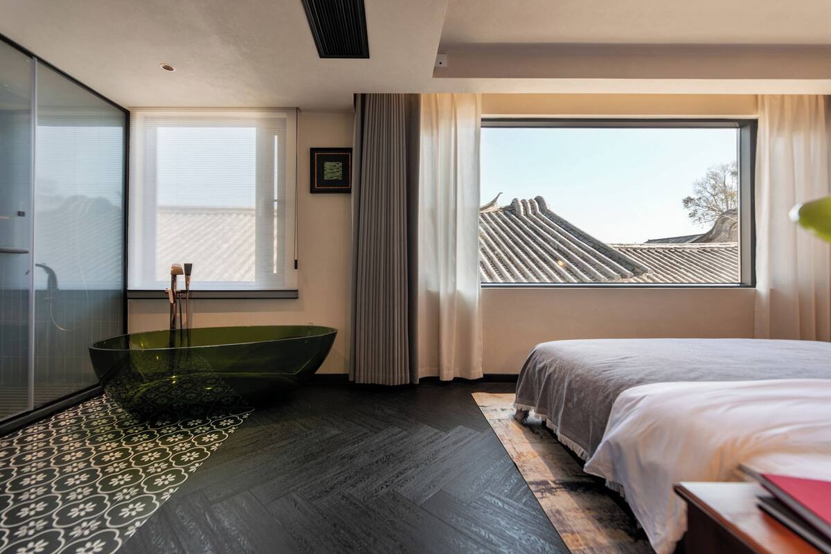 Quarto moderno com uma janela grande, uma banheira verde e uma cama com edredom branco.