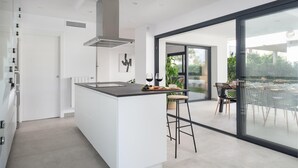 A modern kitchen with a central island, a dining area with a glass door leading to an outdoor patio, and a large window providing a view of the outdoors. - Ca Na Cati (barcarÉs) - Villa With Private Pool (Es Barcarès (alcúdia), Illes Balears)