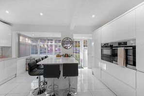 A modern kitchen with white cabinetry, a central island with bar stools, a large window with a view, and built-in appliances. - Family-Friendly 4-Bed near Parks & Transport Links (Great Barr)