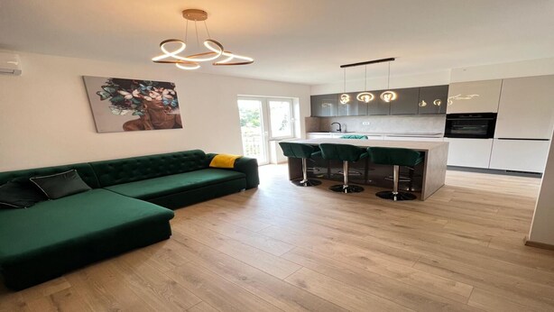 A modern living room with a green sectional sofa, a kitchen with white cabinets, and a bar area with stools. - Apartments Kalinić (Janjina)