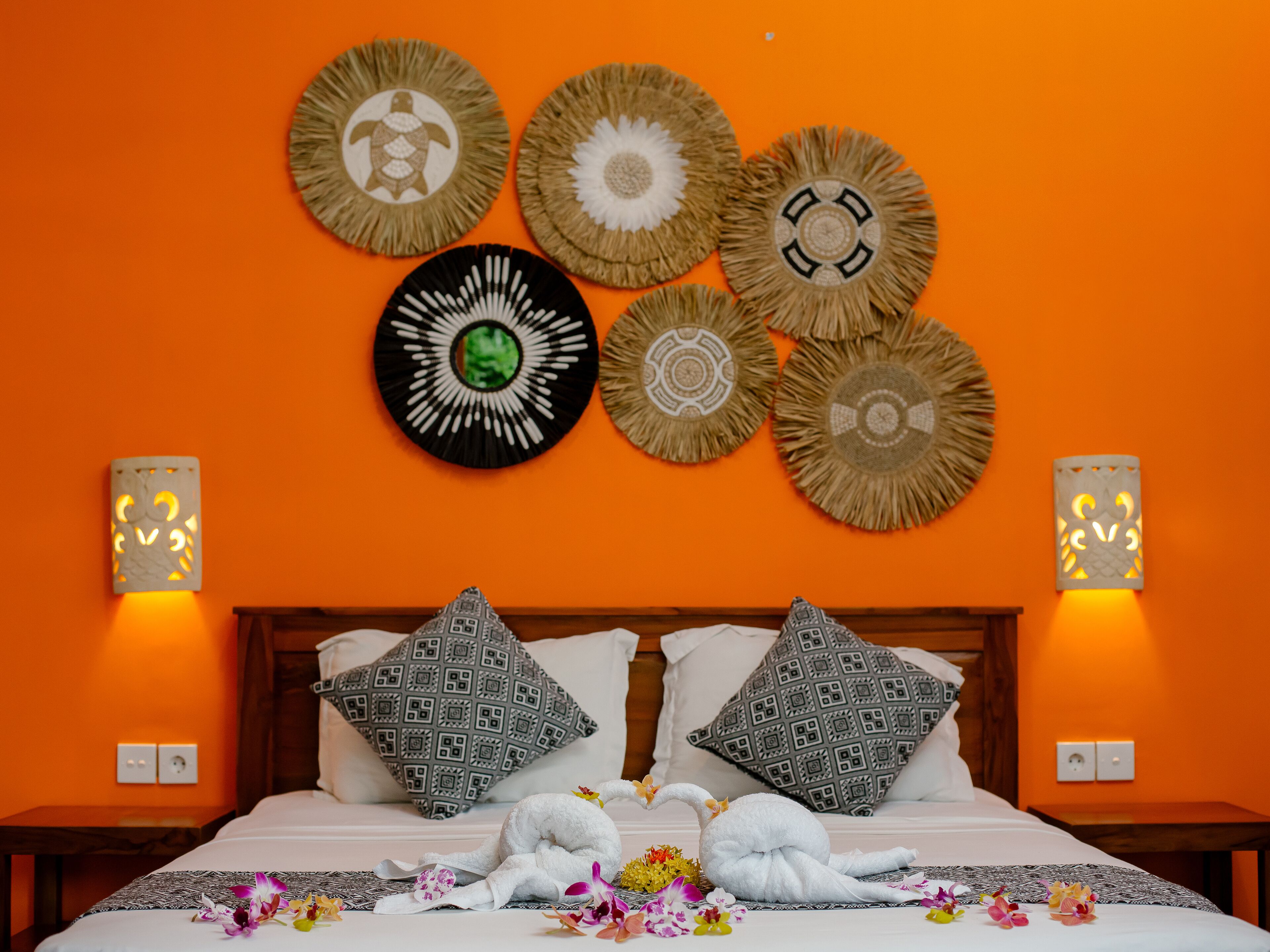 A hotel room with an orange wall, a bed with patterned pillows, decorative wall hangings, and two wall-mounted lamps.