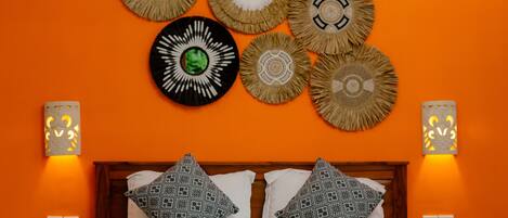 A hotel room with an orange wall, a bed with patterned pillows, decorative wall hangings, and two wall-mounted lamps.
