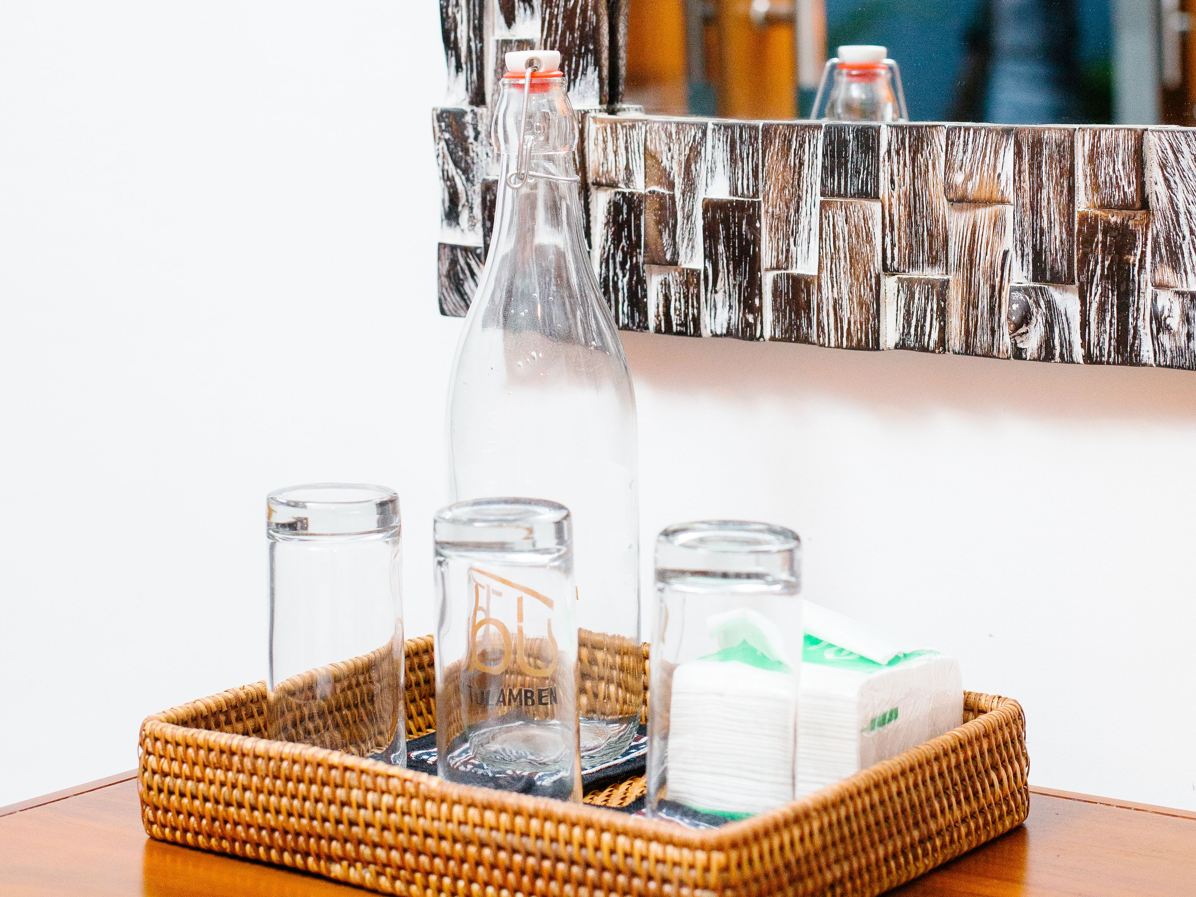 A wicker tray with glass bottles, a glass, and toilet paper on a wooden table.