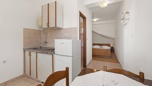 A compact kitchen with a refrigerator, sink, and wooden cabinets. A dining table with chairs is in the foreground. A bedroom with a bed and nightstand is visible in the background.