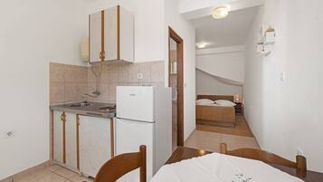 Eine kleine Küche mit Kühlschrank, Spüle und Holzausbau. Im Vordergrund steht ein Esstisch mit Stühlen. Im Hintergrund ist ein Schlafzimmer mit Bett und Nachttisch zu sehen.