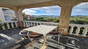 A covered outdoor seating area with a table and chairs, a white balustrade, and a view of greenery and buildings in the distance. - Studio in Tribunj near Bristak Beach (Tribunj)