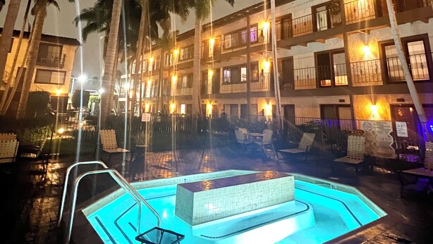 A nighttime pool area with a blue-lit pool, palm trees, and a multi-story building with balconies. - Resort Style Home near Beaches & Disneyland (Fountain Valley)