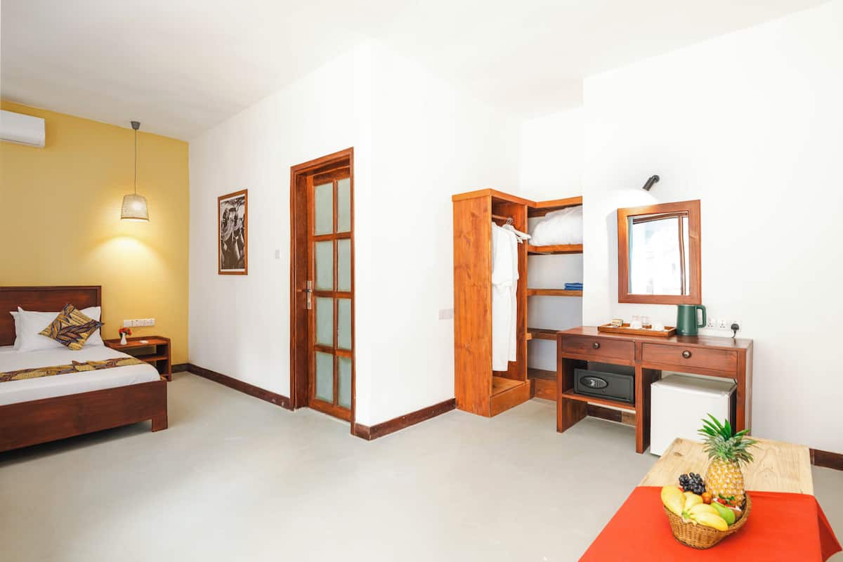 Habitación de hotel con cama, escritorio, refrigerador pequeño, frutero y puerta que da a otra habitación.