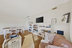 A modern living room with a white sofa, a wooden dining table, a white chair, a bookshelf, and a television.