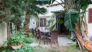 A patio with a table and chairs, surrounded by lush greenery and a building with a tiled roof.