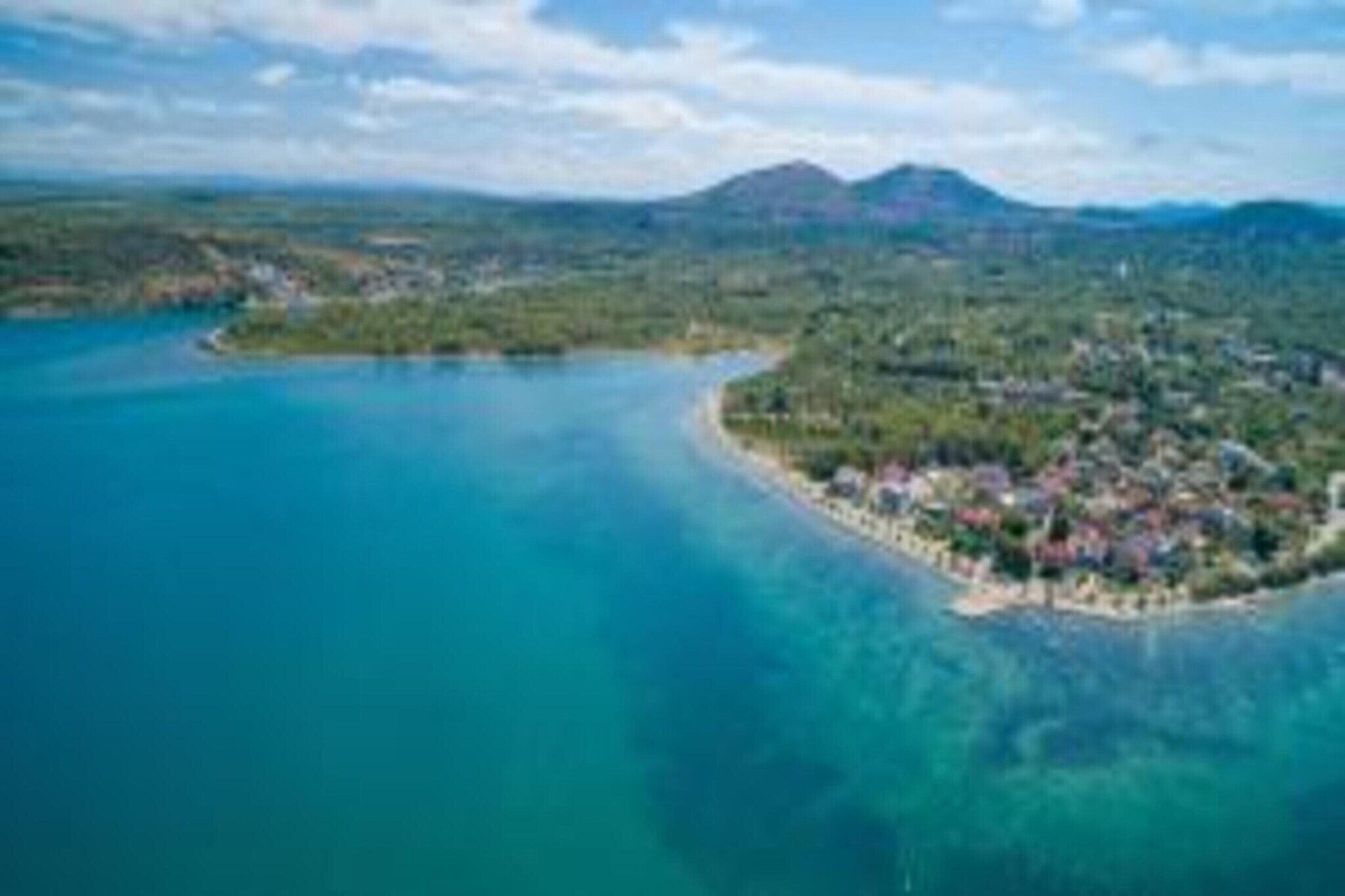 An aerial view of a coastal area with clear blue waters, a small peninsula with buildings, and a mountain range in the background.