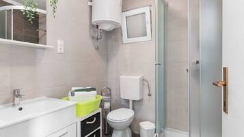 A modern bathroom with a white sink, toilet, and shower. There is a green trash bin and a white basket on the sink.
