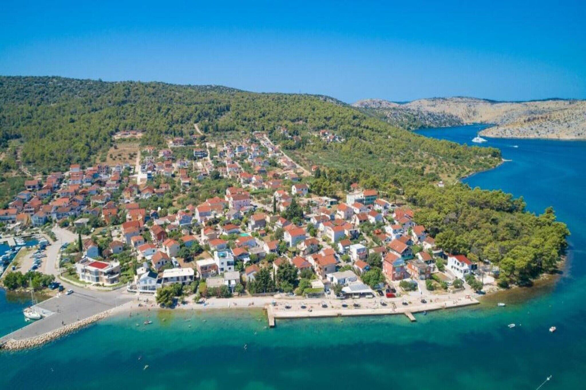An aerial view of a coastal village with houses, a marina, and a clear blue sea.