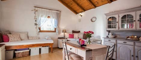 A cozy room with a wooden ceiling, a bed with a white canopy, a dining table with chairs, and a kitchen area with cabinets.