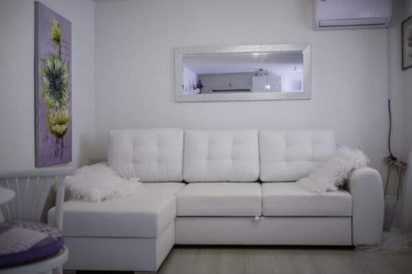Un canapé blanc surmonté d’une couverture moelleuse également blanche, une petite table ronde avec un coussin violet, et un miroir encadré accroché au mur.