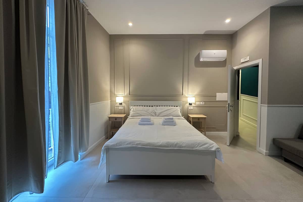Ein Hotelzimmer mit einem großen Bett, zwei Nachttischen, einer Wand-Klimaanlage und einer Tür zu einem anderen Raum.