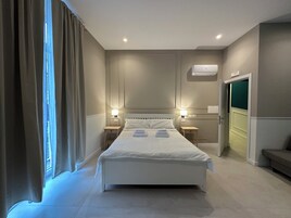 Una camera d'albergo con un letto grande, due comodini, un condizionatore d'aria a parete e una porta che dà su un'altra stanza.