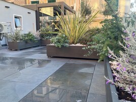 Un cortile con pavimento piastrellato, fioriere con diverse piante e una facciata di edificio moderna.
