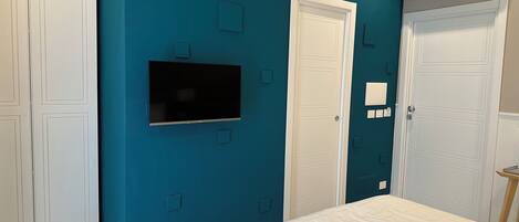 A bedroom with teal walls, a flat-screen TV mounted above a door, white bedding, and a stack of folded towels.