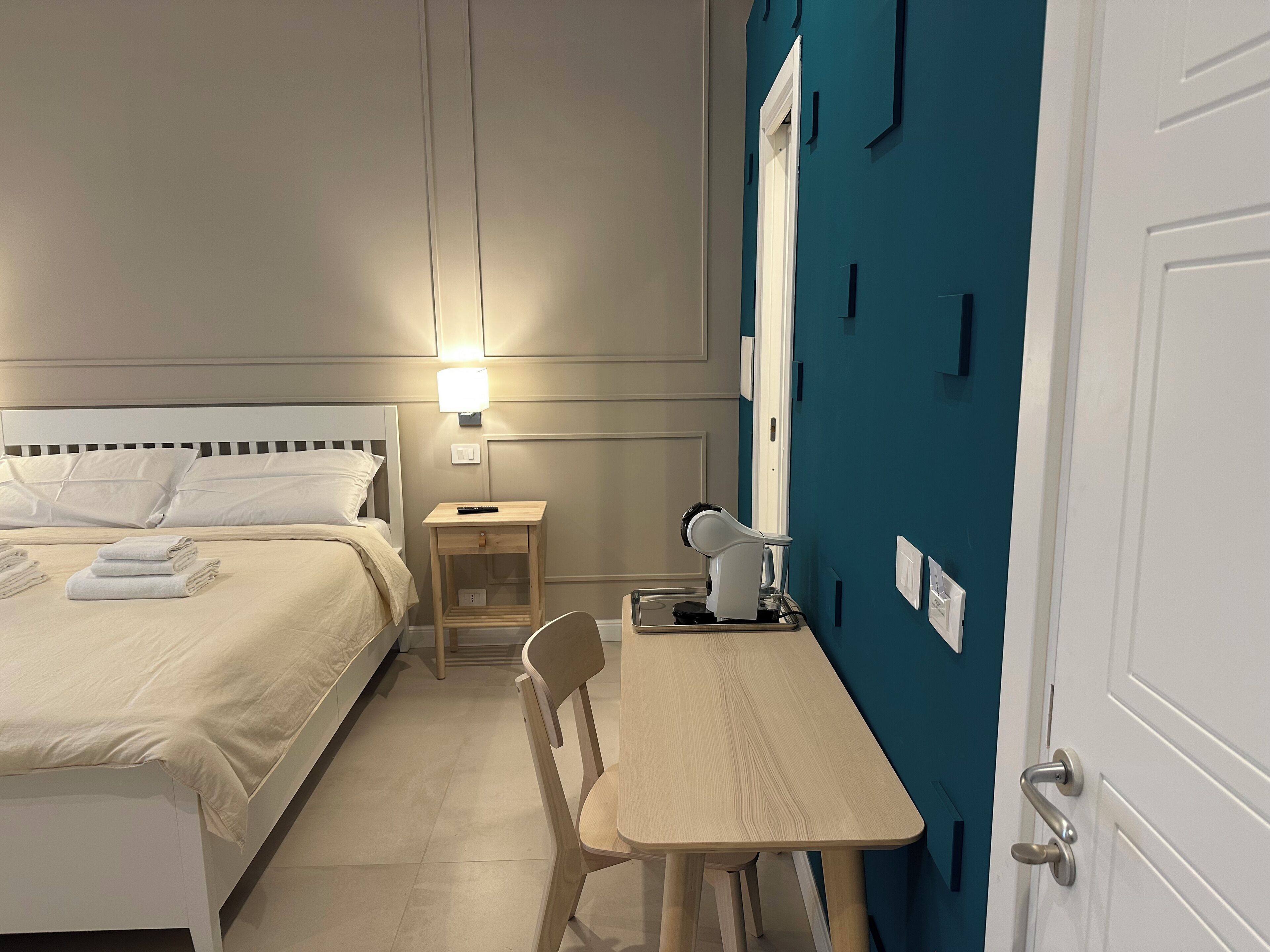Ein ordentlich eingerichtetes Hotelzimmer mit Bett, Nachttisch, Stuhl, Schreibtisch und einer Tür.