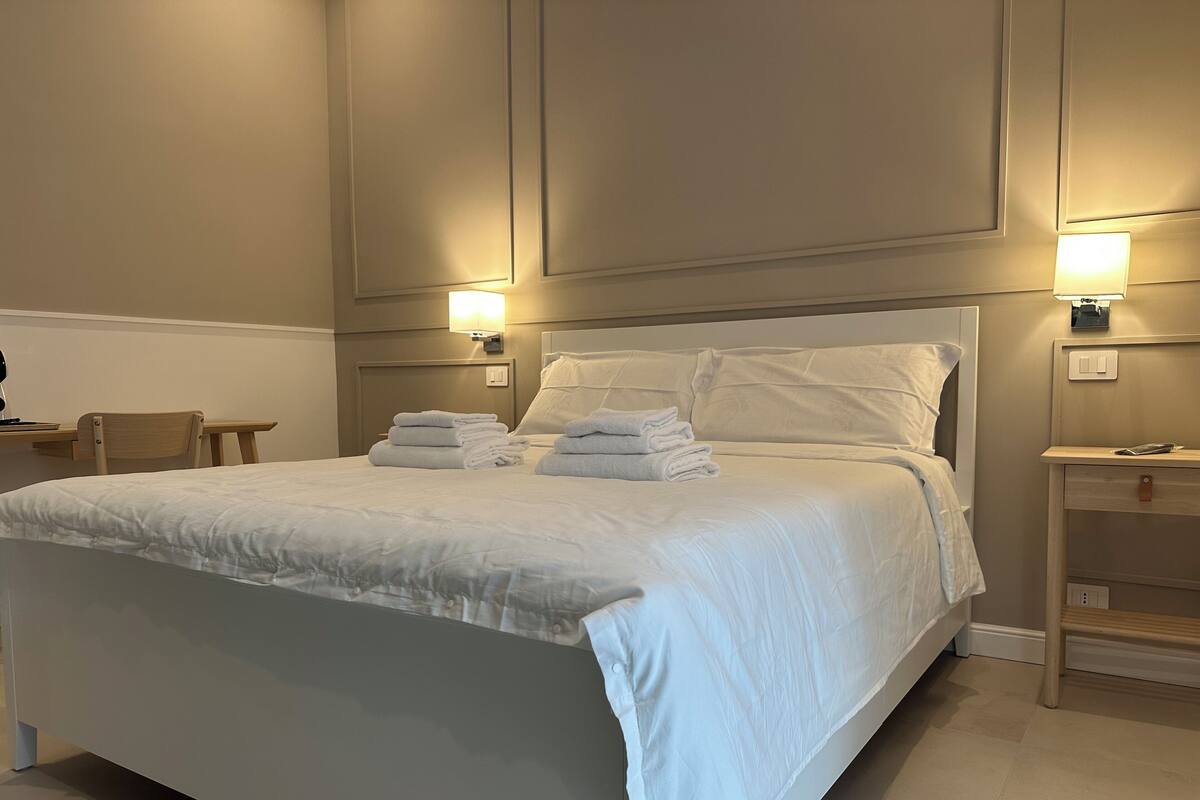 Ein ordentlich gemachtes Bett mit weißen Bettwäsche und Handtüchern, ein Nachttisch mit Lampe sowie ein Beistelltischchen mit Telefon.