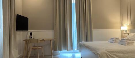 A hotel room with a bed, a desk with a chair, a television, and a window with curtains.