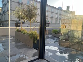 Un edificio moderno con ingresso in vetro, area esterna con posti a sedere e piante, e pavimento piastrellato.