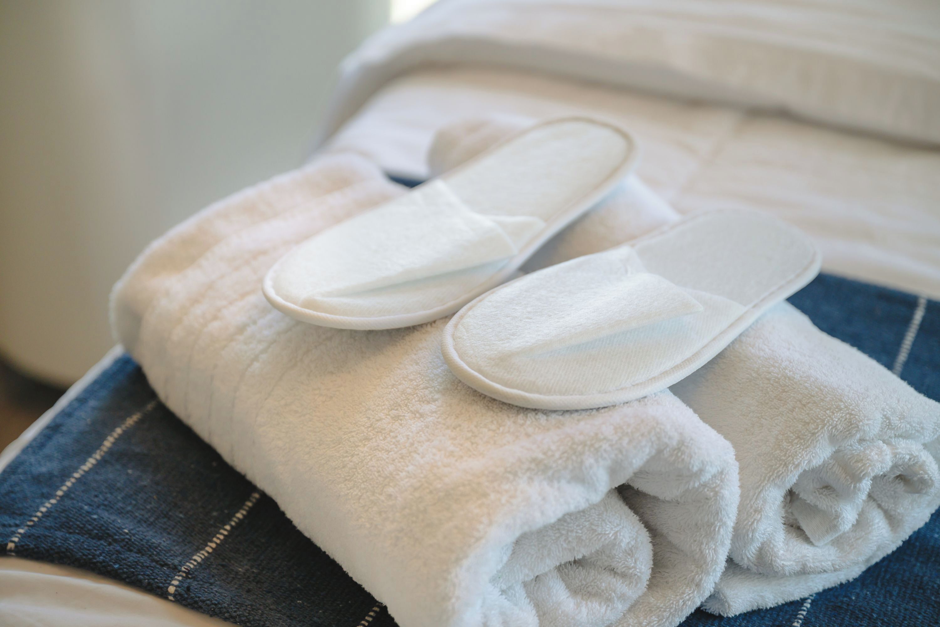 A neatly folded white towel and a pair of white slippers on a bed with a blue striped pillow.