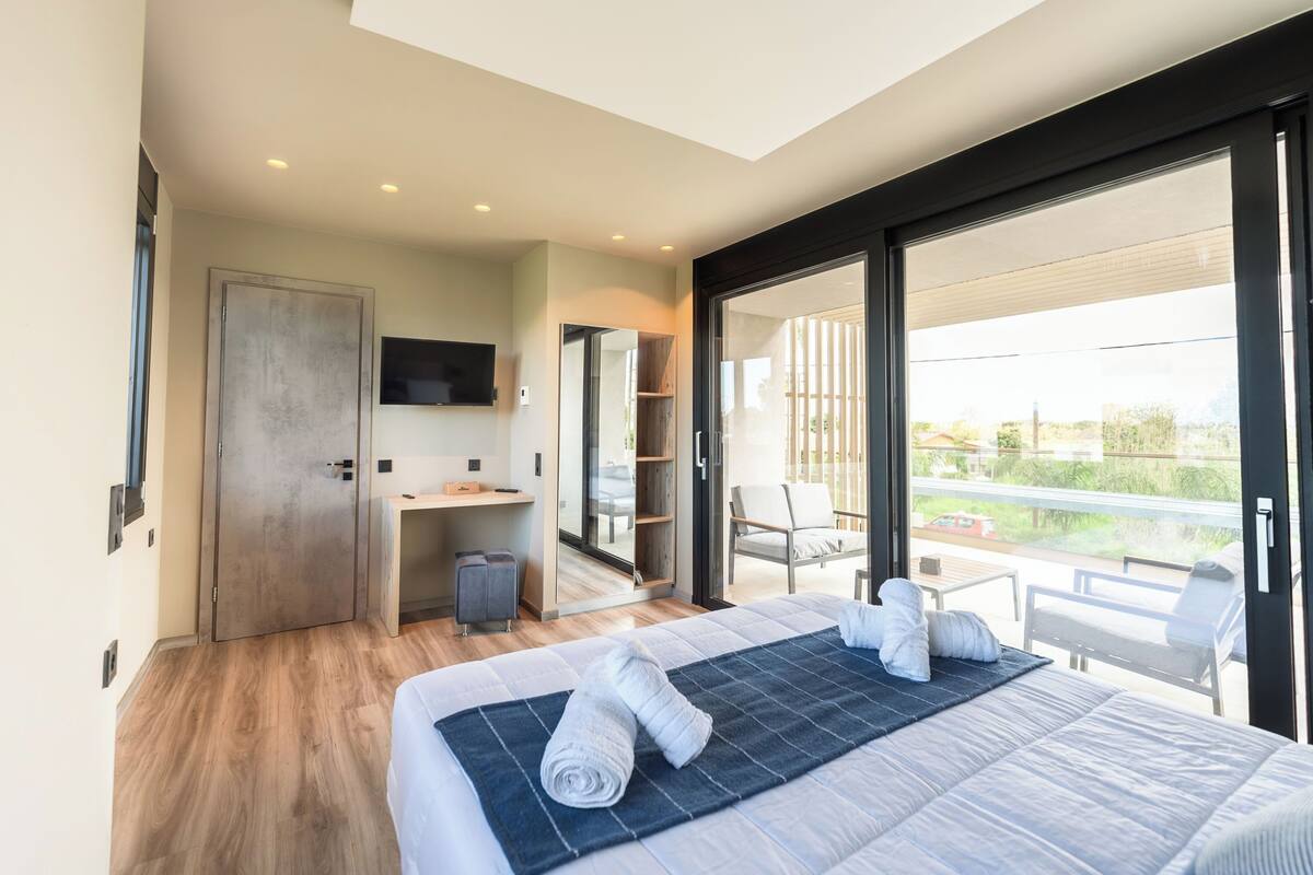 A modern hotel room with a large bed, a desk, and a balcony with outdoor seating.